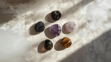 Five protective crystals — obsidian, tourmaline, amethyst, smoky quartz, and tiger’s eye on neutral background.